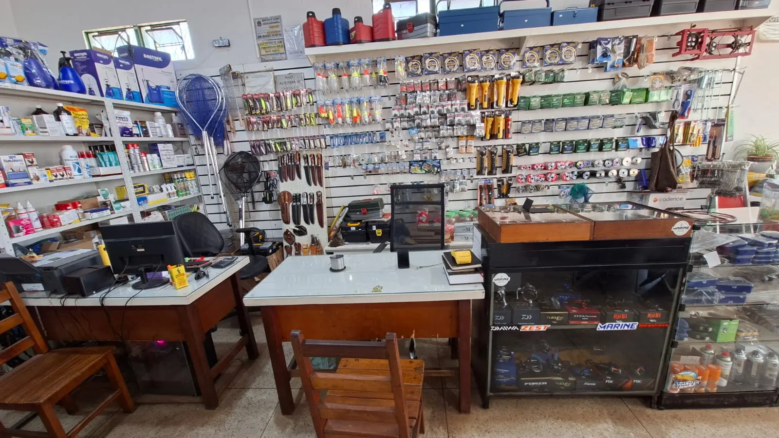 Balcão de atendimento do setor de pesca, com vitrines de molinetes e carretilhas, parede de anzóis, iscas e linhas, e o computador do caixa sobre a mesa de madeira.