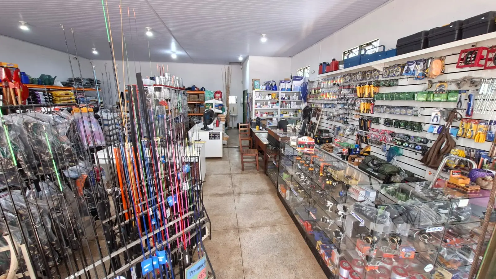 Corredor de pesca com dezenas de varas em pé no expositor à esquerda e vitrines de vidro com molinetes e acessórios à direita.