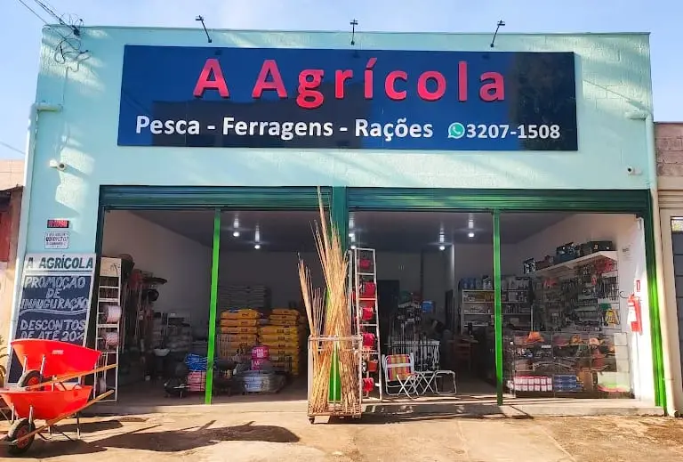 Fachada da A Agrícola: parede verde-água e letreiro azul com o nome da loja em vermelho, a linha 'Pesca - Ferragens - Rações' e o telefone 3207-1508. Os dois portões verdes estão abertos, com um carrinho de mão vermelho na calçada.